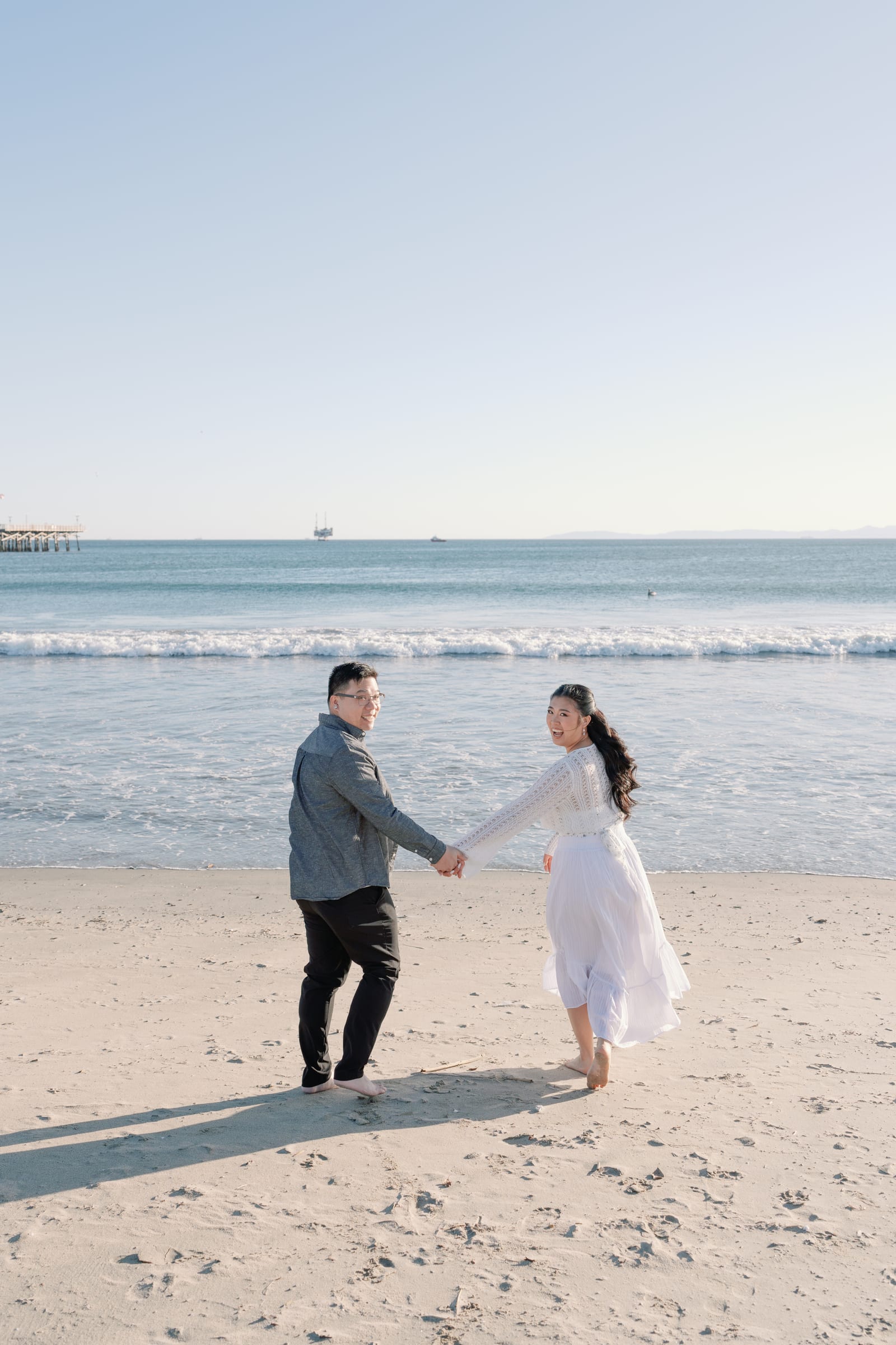 Christine and Thomas holding hands on the beach, glancing back at the camera.